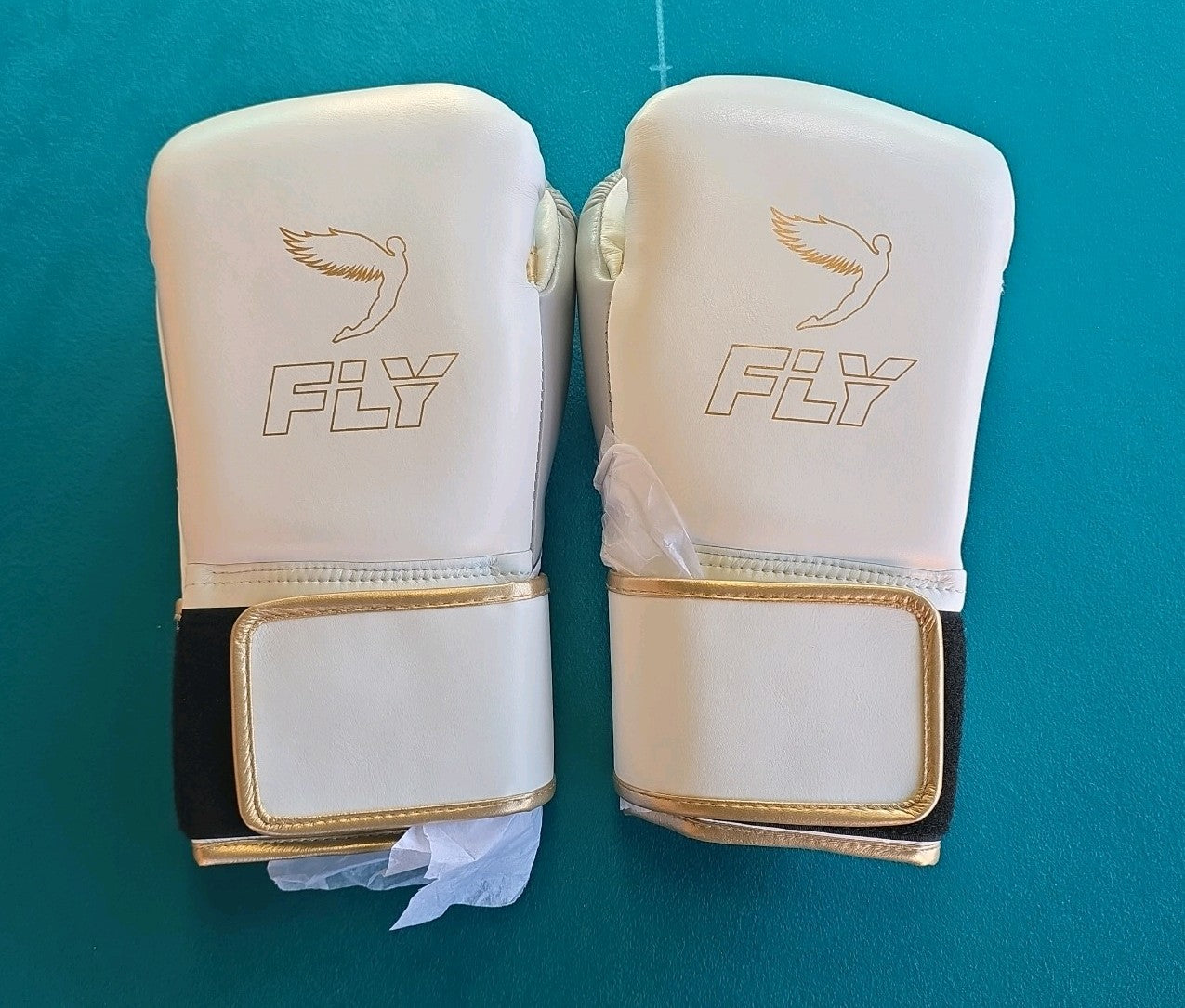 Pair of white boxing gloves with gold trim and FLY logo on teal background, with Velcro wrist straps.