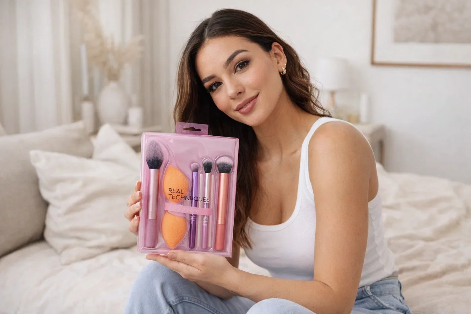 Smiling woman on a bed holds a pink Real Techniques makeup brush set labeled Hot Seller.